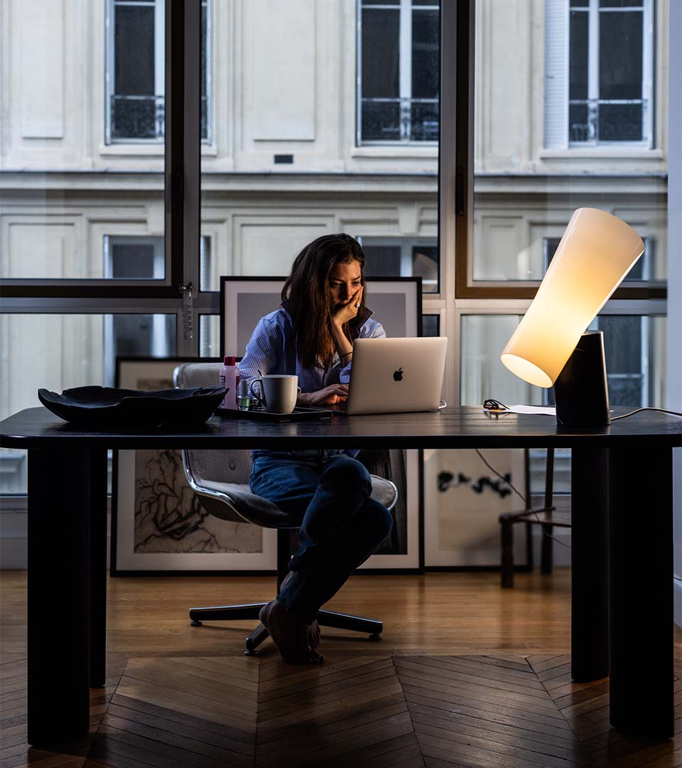 Nő laptopon dolgozik egy íróasztalnál, világító asztali lámpa mellett