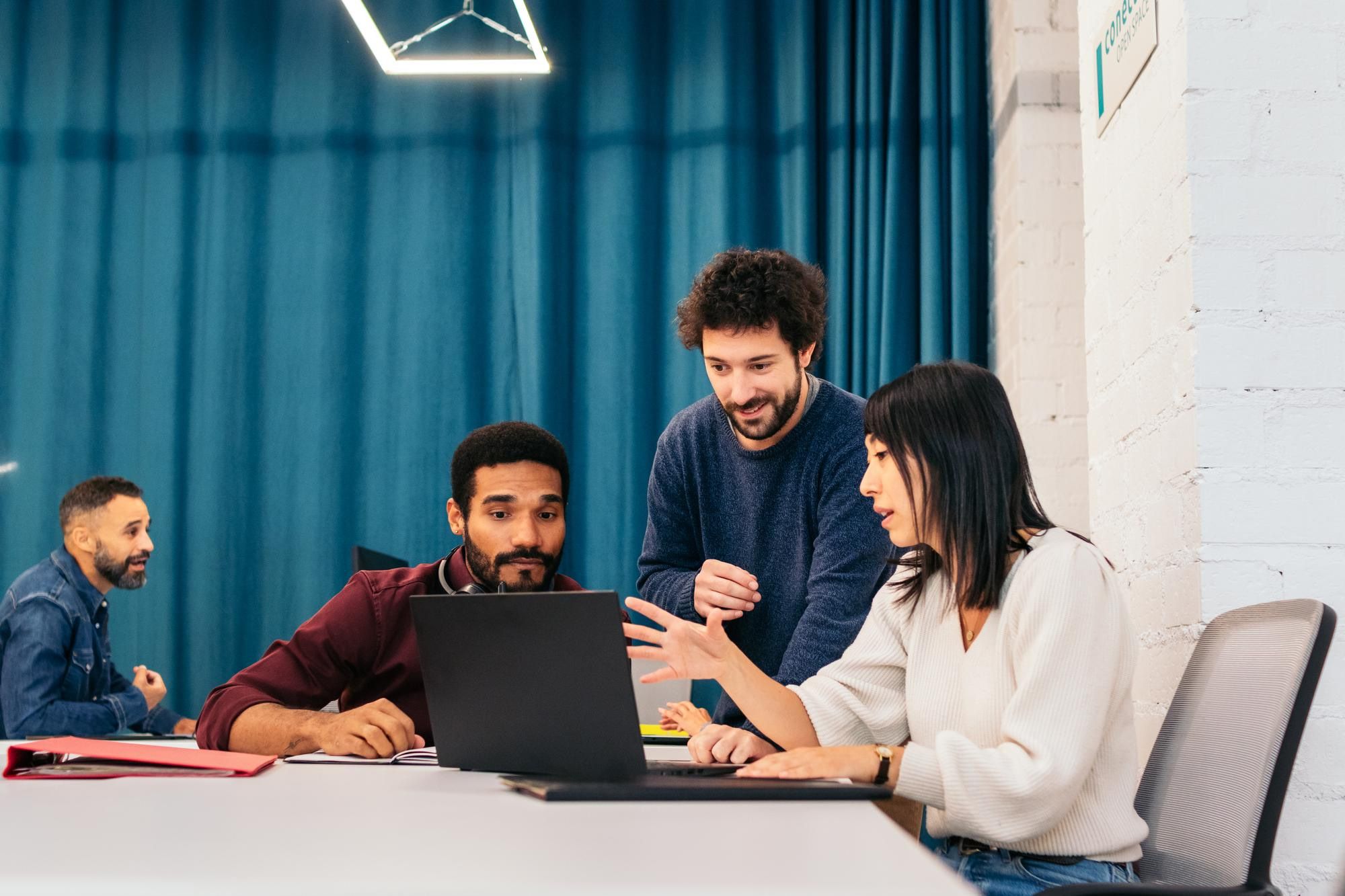 Four colleagues around a table in an office, talking through work on a laptop.