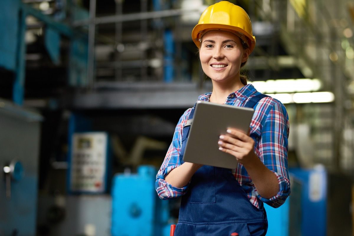 A technician in a hard hat reviewing information on a tablet on a plant floor.