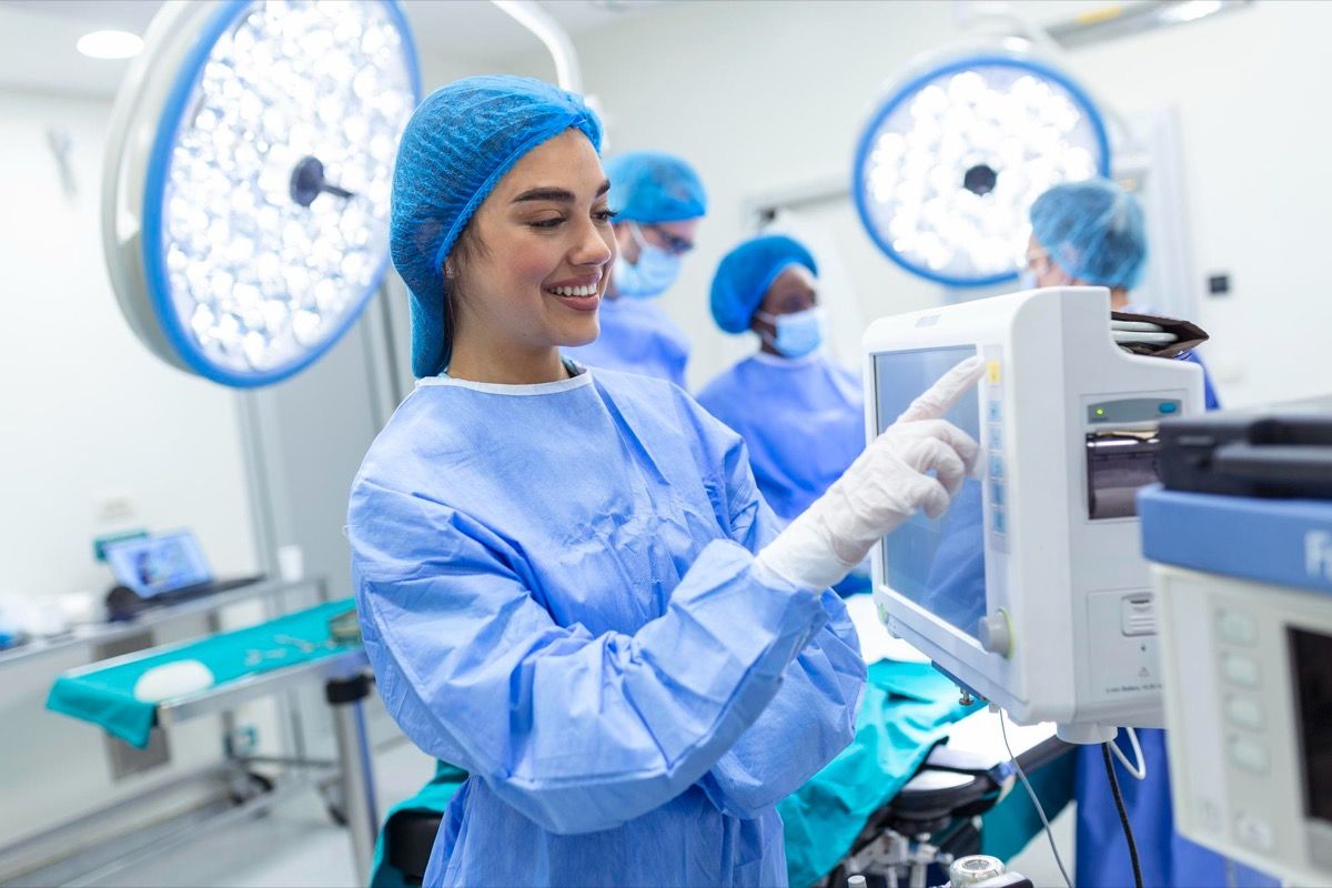 A clinician in surgical scrubs adjusting settings on a patient monitor in an operating theatre.