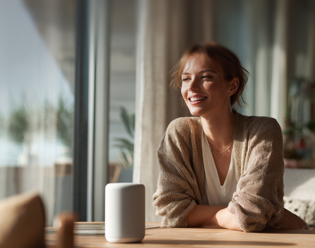 A smiling woman sitting at a wooden table in a sunlit room, pleasantly interacting with her white smart speaker.