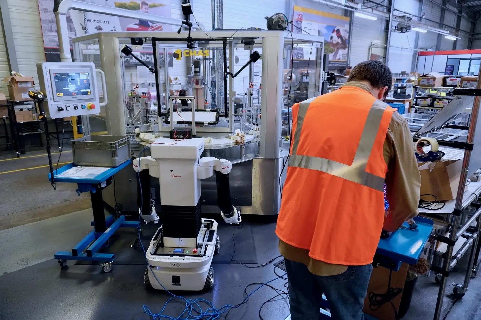 An operator in a hi-vis vest works at a packaging cell beside a humanoid robot