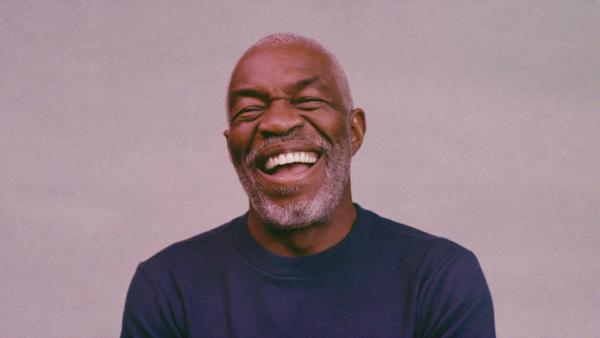 A studio portrait of a man with a short white beard in a plain dark knit, laughing with his eyes crinkled