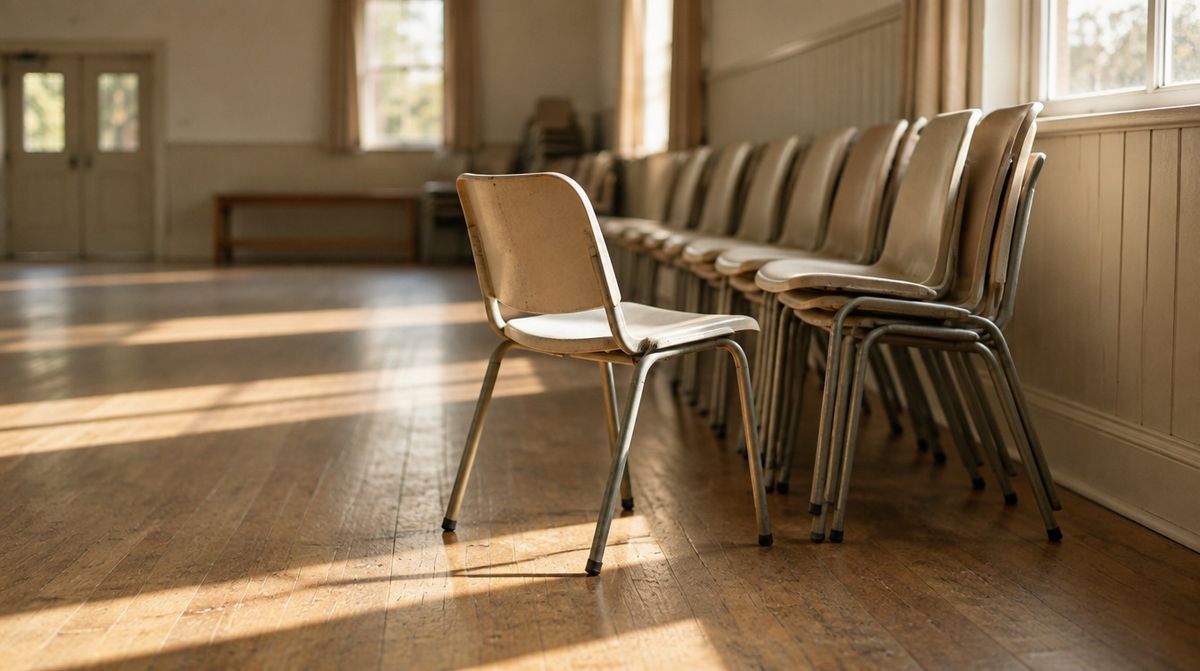 A row of chairs in a community hall, one pulled out from the line, morning light across the floor