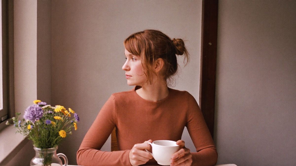 A woman in a rust jumper at a kitchen table, holding a white mug and looking out of the window, a jug of garden flowers beside her