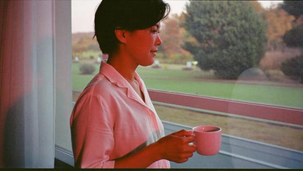 A woman in a plain cream linen shirt standing in profile at a window with a mug, lit from the side
