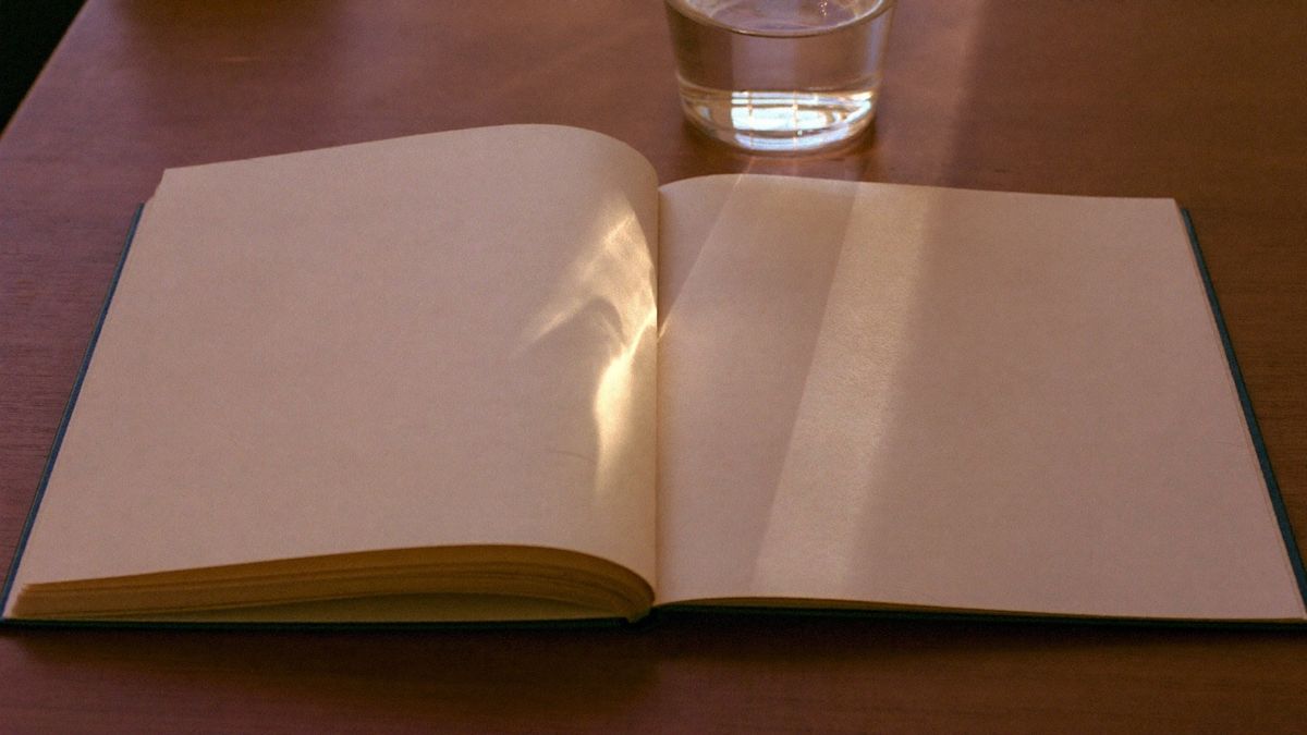 An open notebook with blank cream pages beside a glass of water on a timber table in low golden sun