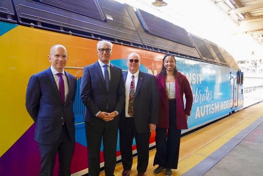 Four people stand in front of an NJ Transit train decorated for Autism Acceptance Month