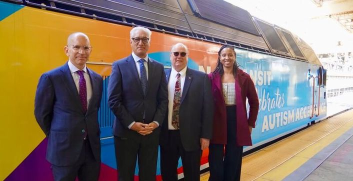 Four people stand in front of an NJ Transit train decorated for Autism Acceptance Month