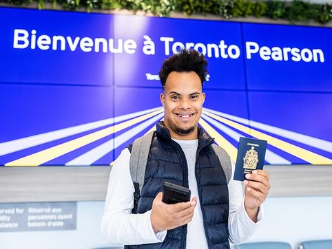 A traveller holds a phone and a passport under a sign reading "Bienvenue à Toronto Pearson"
