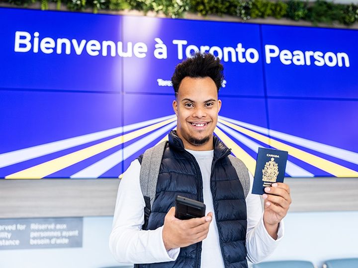 A traveller holds a phone and a passport under a sign reading "Bienvenue à Toronto Pearson"