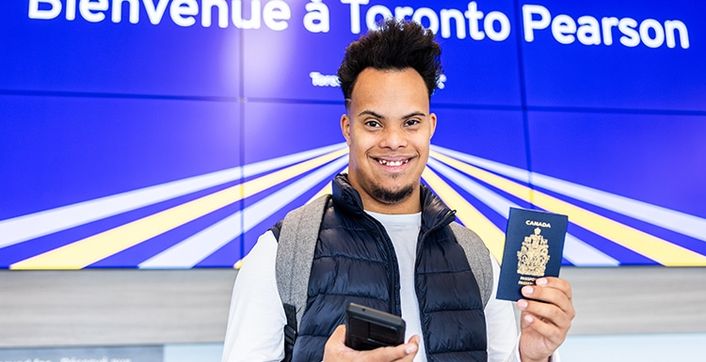 A traveller holds a phone and a passport under a sign reading "Bienvenue à Toronto Pearson"