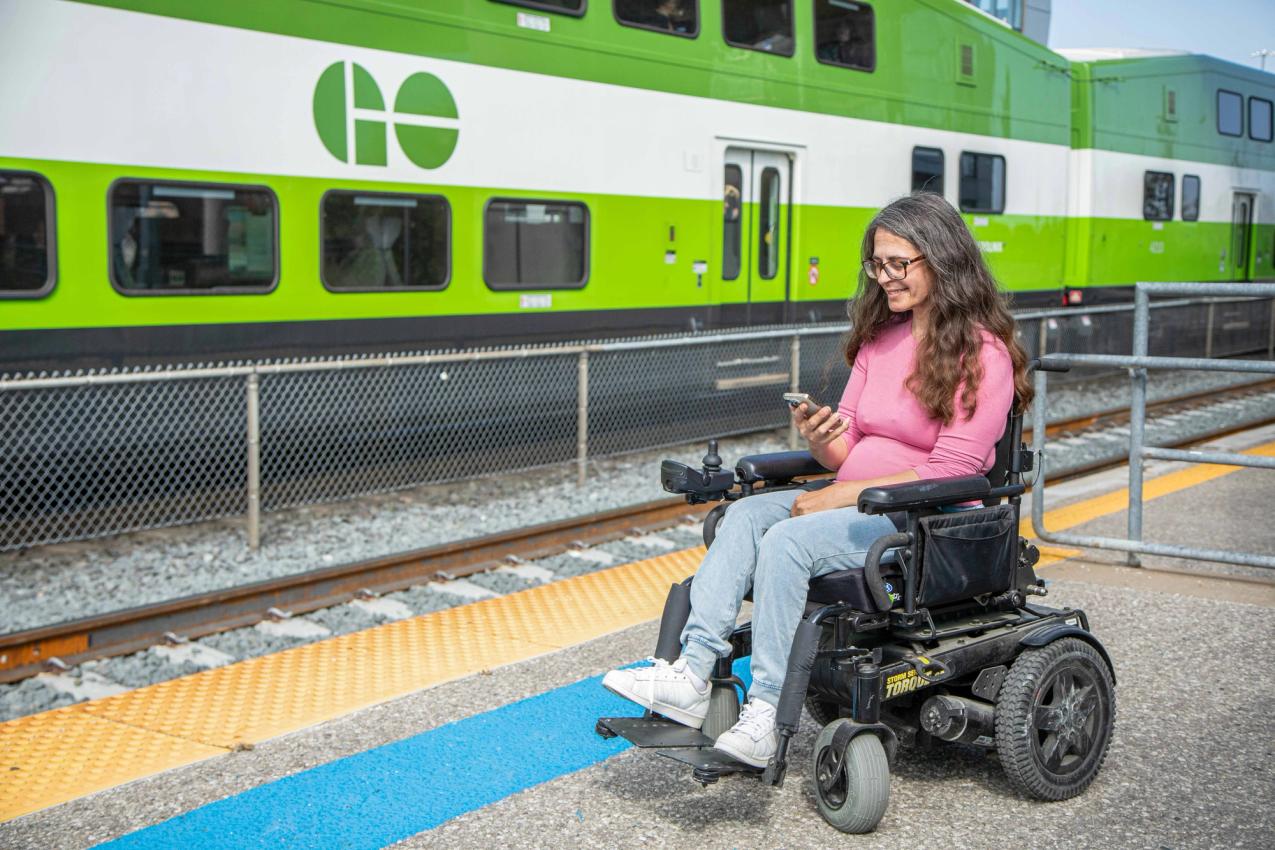 A rider in a power wheelchair uses a phone on a GO train platform
