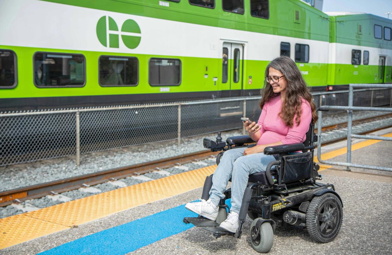 A rider in a power wheelchair uses a phone on a GO train platform