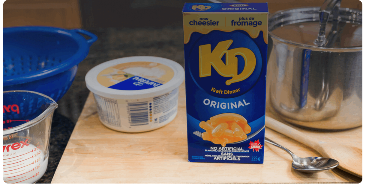 A box of Kraft Dinner with a pot, a colander and a measuring cup