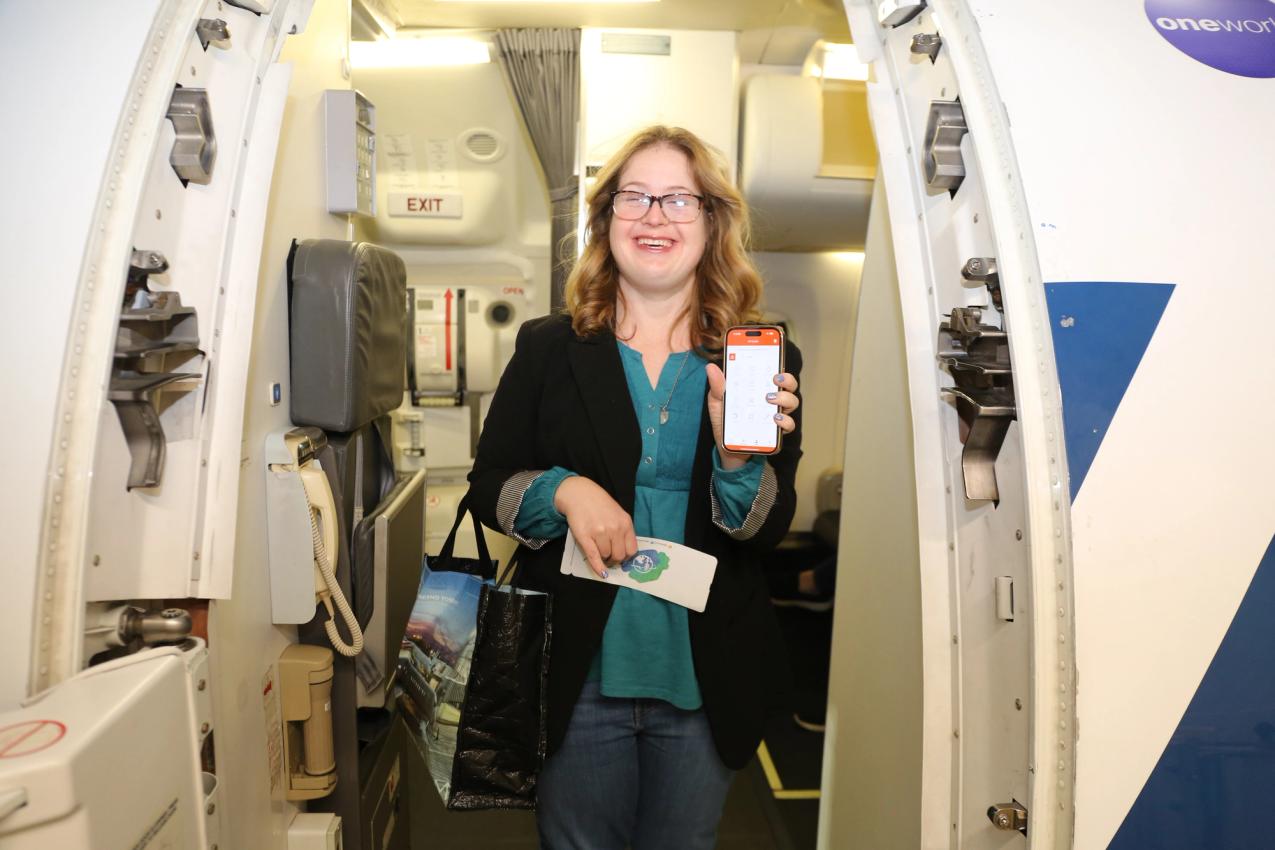 A traveller holding a phone smiles in the doorway of an airplane