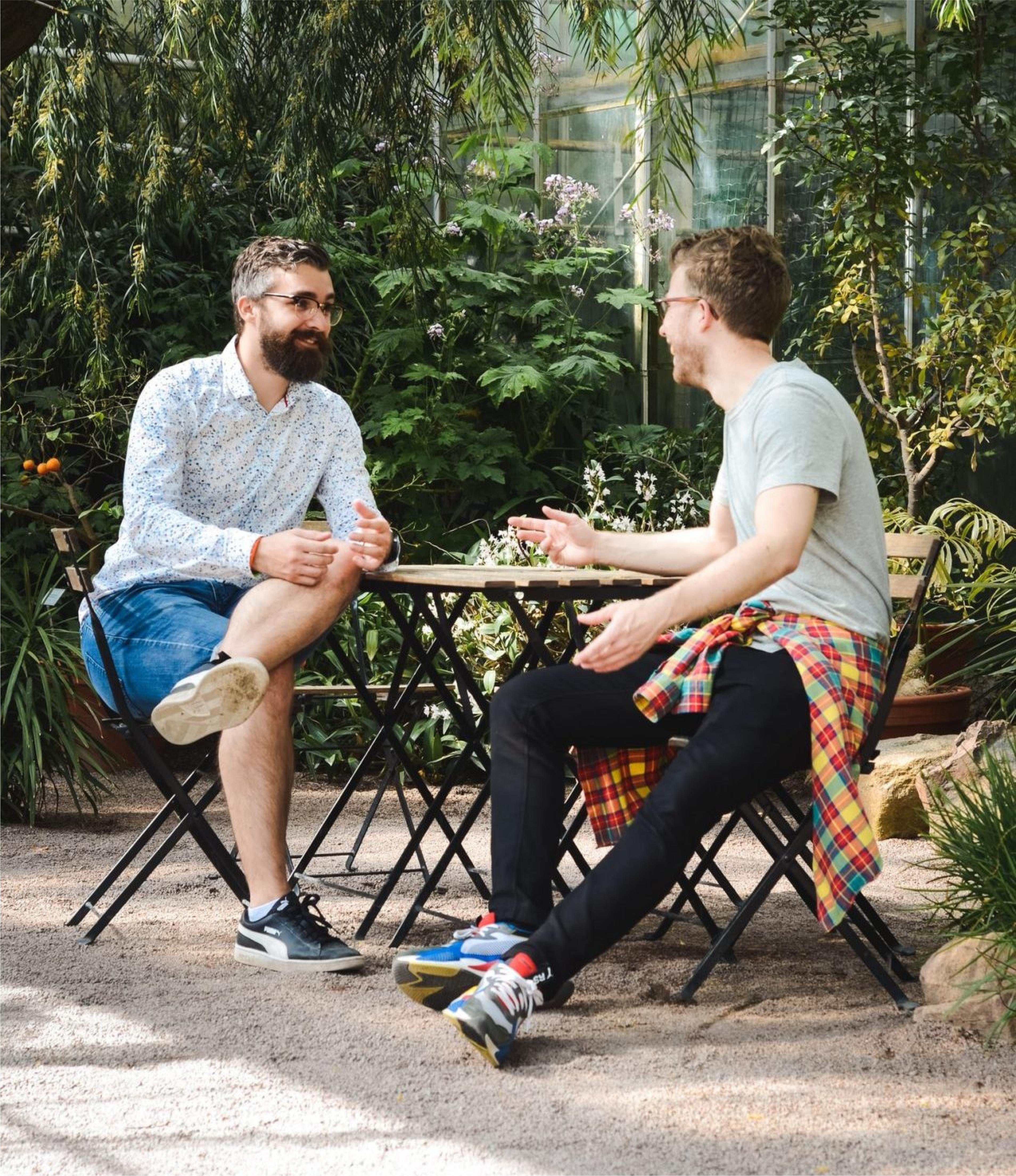 To menn snakker ved et cafébord i grønne omgivelser.