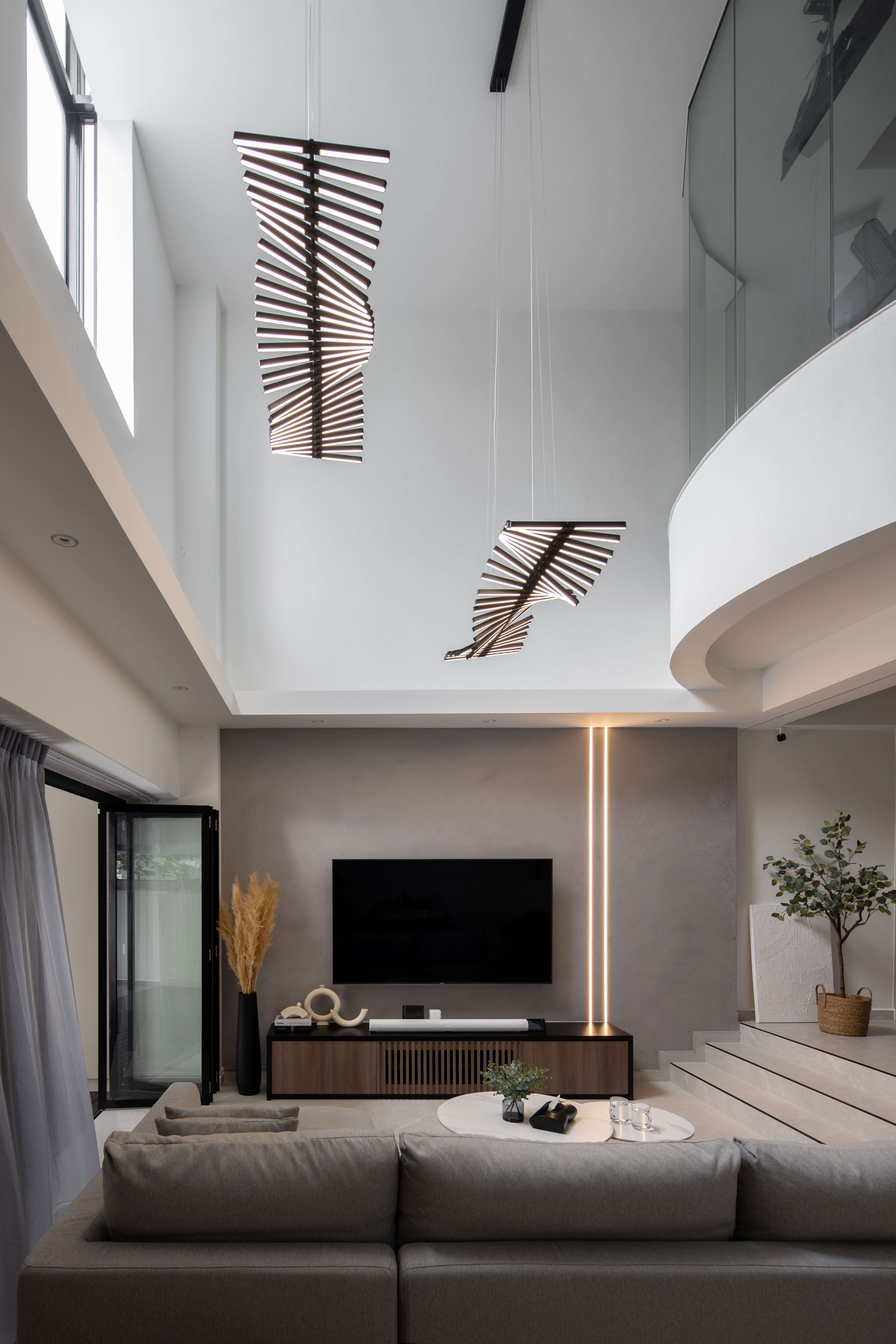 Modern high-ceiling living room with sculptural pendant lights, neutral-toned sofa set, and elegant ambient lighting along the stairs and entertainment area.