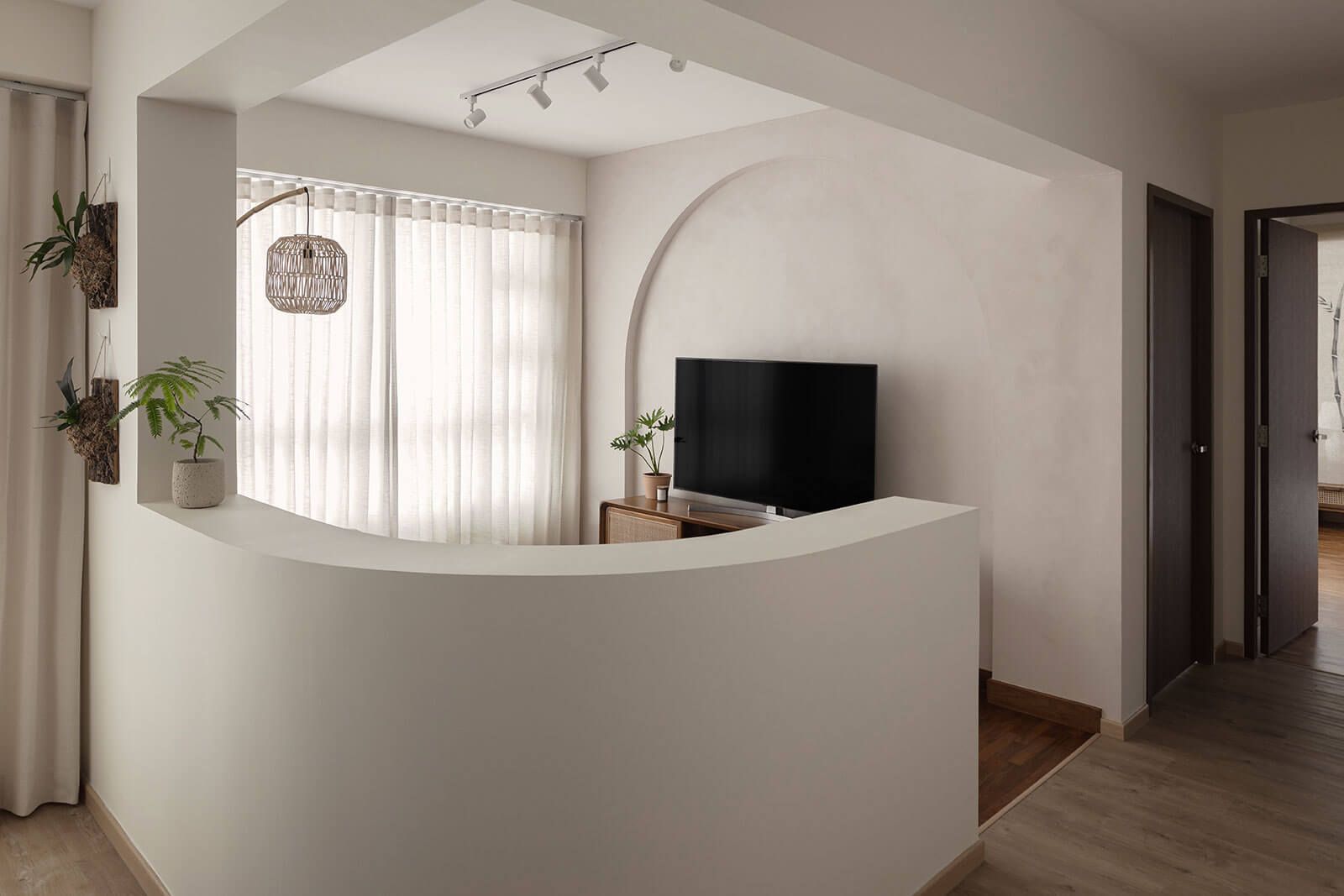 Modern minimalist TV wall design featuring a curved wall niche with a flat-screen TV, wooden console, and hanging plants against sheer curtains and wooden flooring.