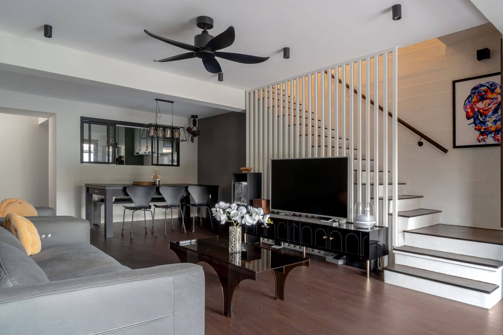 Open-plan living space with a sleek TV console against a wooden slatted partition, complemented by a contemporary dining area, artistic decor, and a statement staircase in a stylish home.