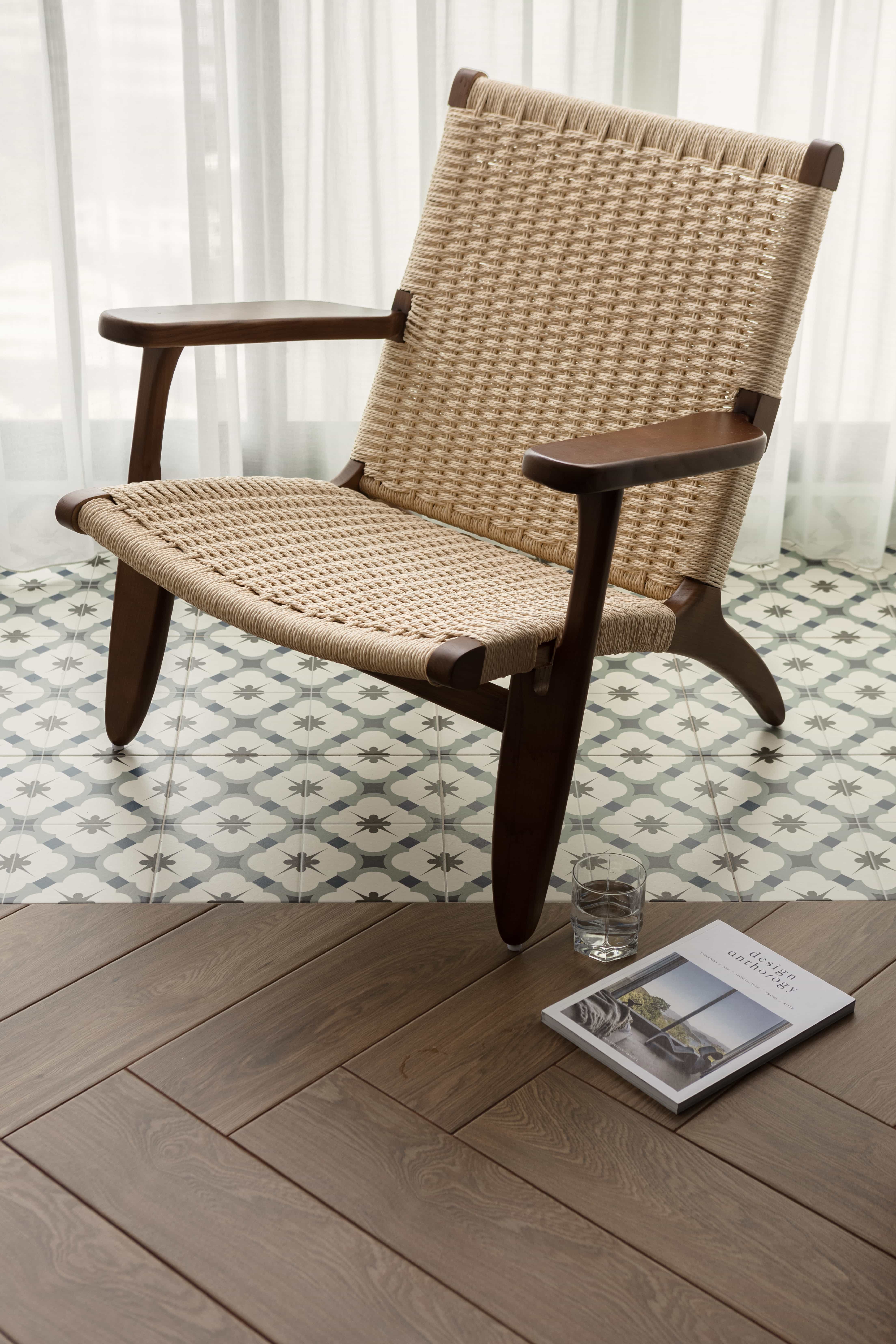 Woven chair with wooden frame on patterned floor tiles next to a magazine and glass of water.