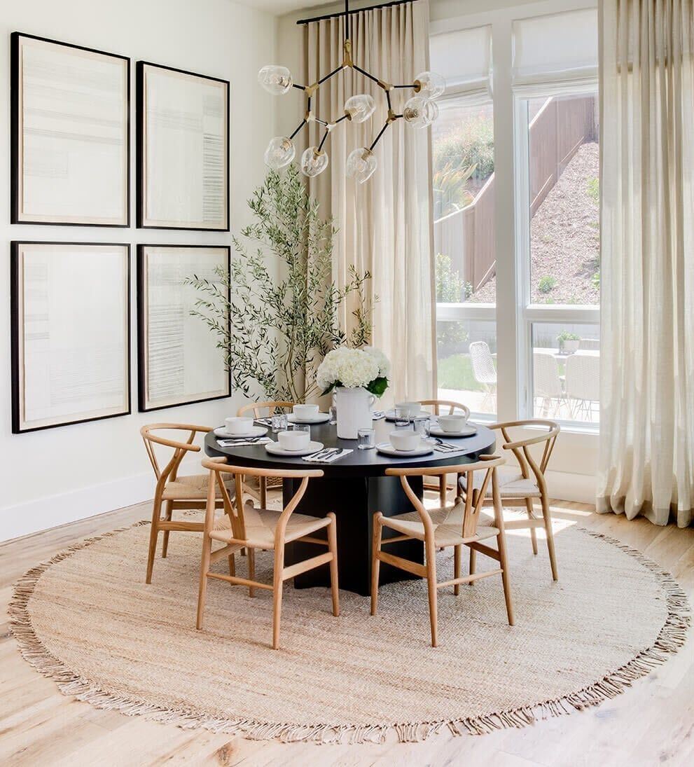 A round jute rug in a dining room with natural wooden chairs and a black round dining table, framed by large windows and wall decor.