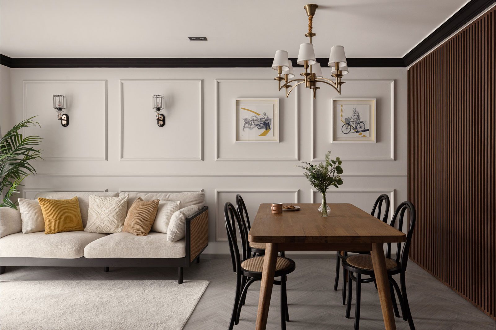 A living room with wainscoted walls, a neutral-toned sofa with yellow cushions, black dining chairs, and a wooden dining table, blending classic vintage and modern interior design elements.