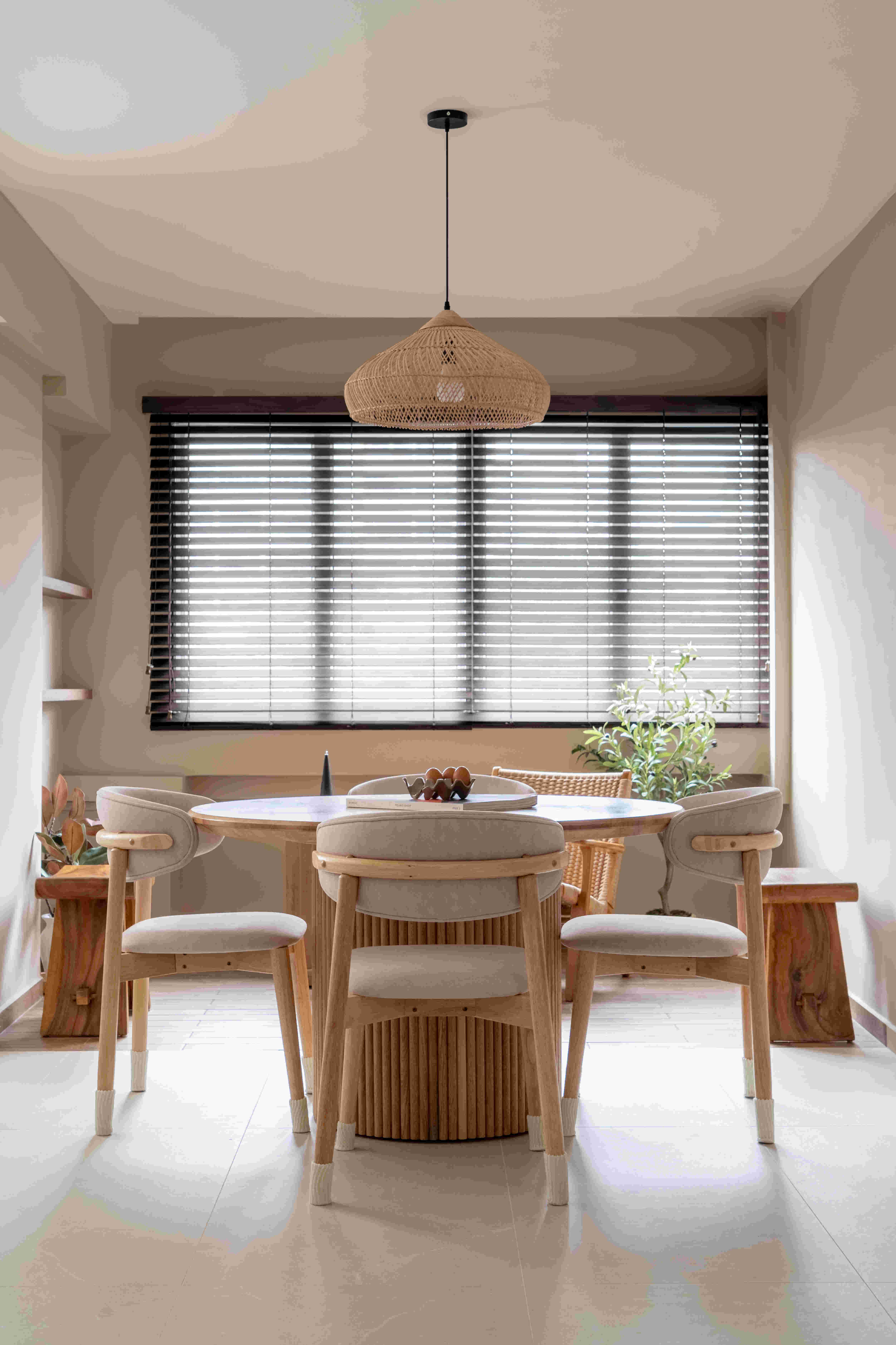 Inviting dining corner with woven rattan pendant, round timber table, sculpted chairs, and potted greenery framed by wide window blinds.