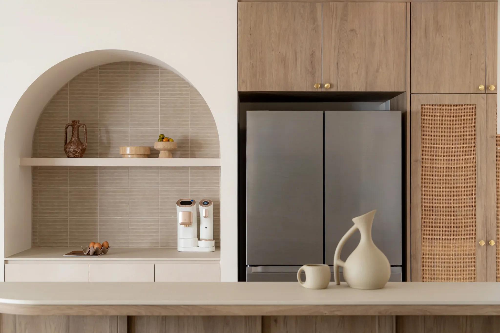 Kitchen with arched alcove shelf, slim beige tiles, stainless French-door fridge, rattan-panel cabinet and brass knobs.
