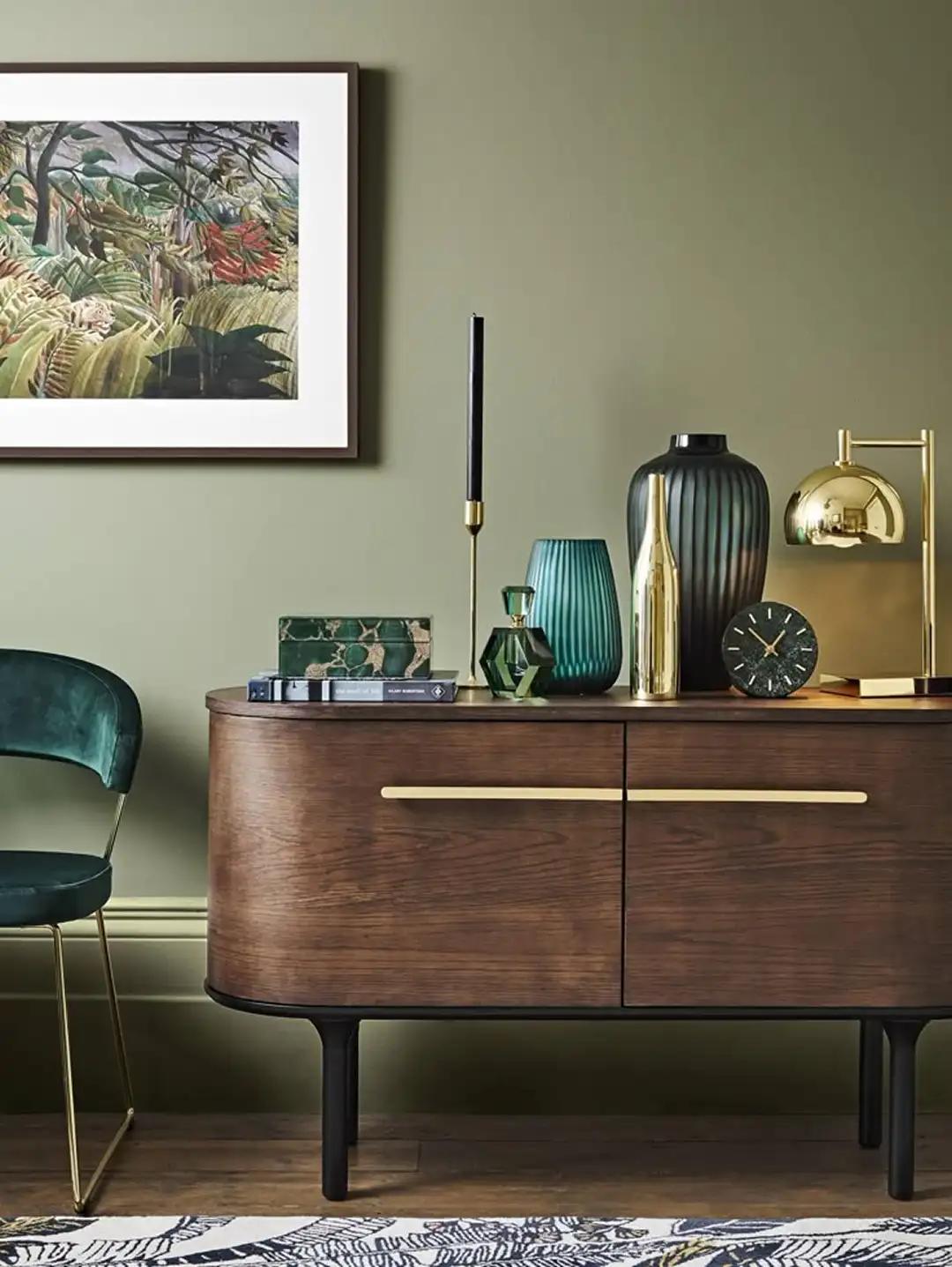 Walnut sideboard against olive wall with brass lamp and green glass accents; modern take on vintage, dodol-toned warmth.