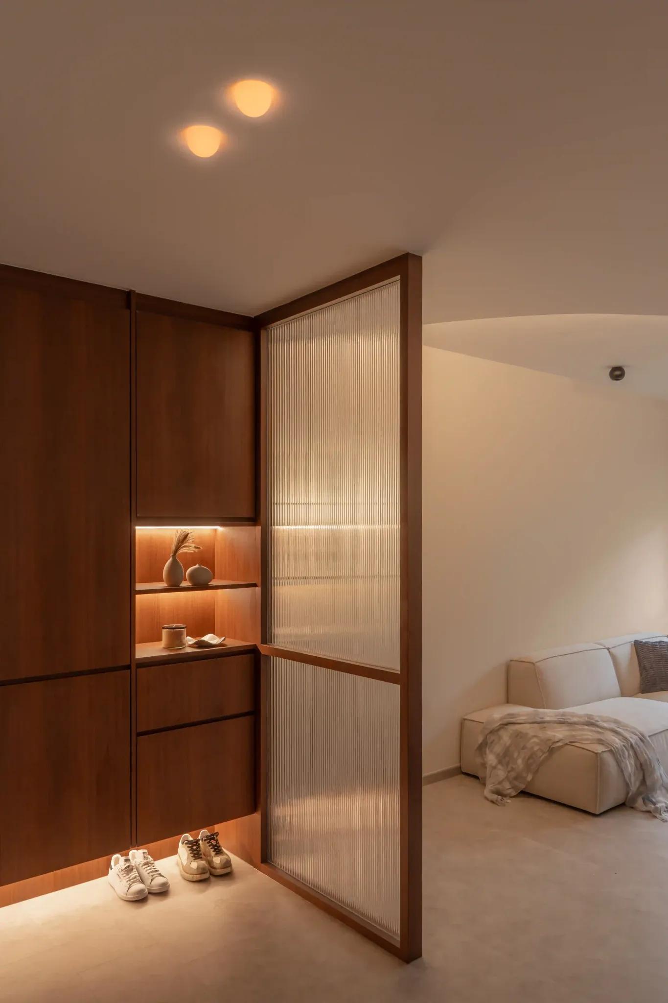 Entryway shoe cabinet in warm wood with reeded-glass screen, lit display shelves, and concealed storage by the living area.