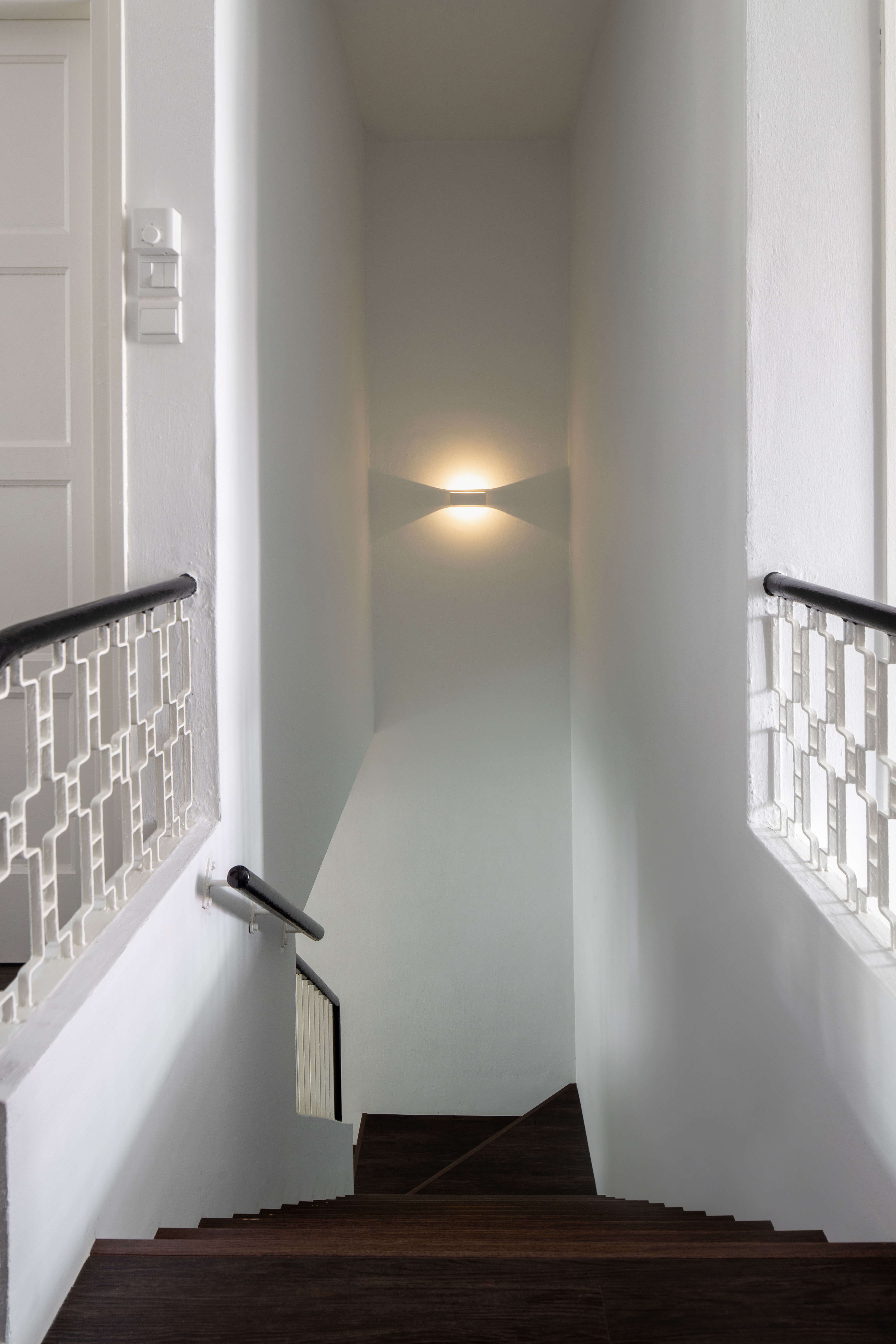 Stairway in a bungalow with decorative wrought iron balustrade and wood railing, illuminated by a warm wall sconce light casting a soft glow in a narrow hallway.