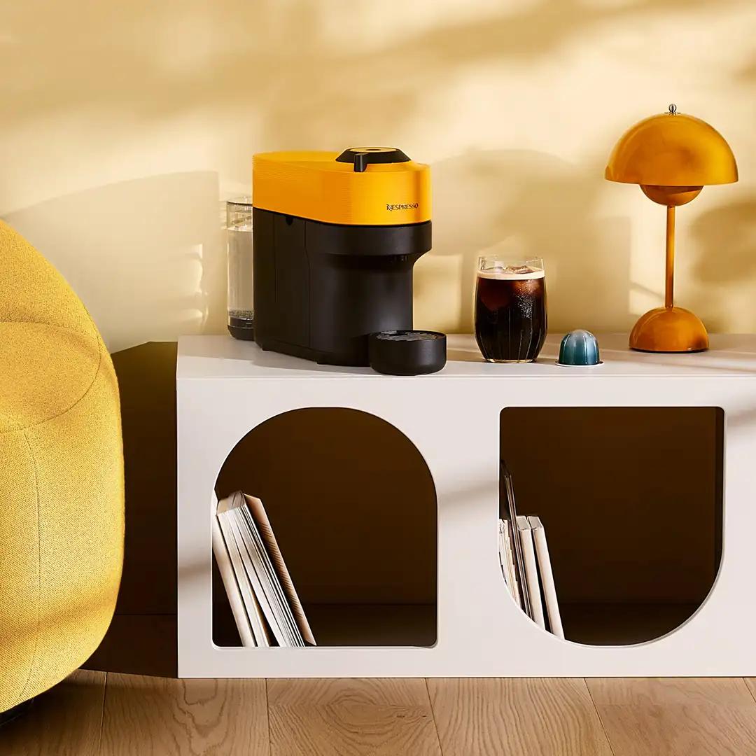 Mango-yellow coffee machine and lamp on white arched shelf beside a soft yellow sofa; cheerful morning nook.