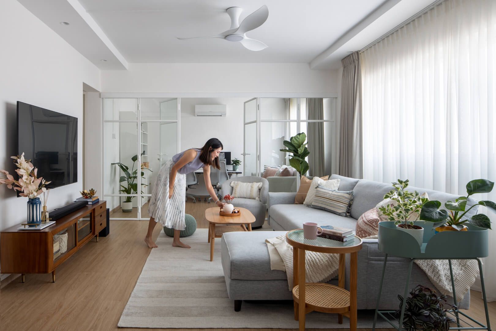 Bright and airy living room with a woman arranging decor, showcasing a grey sofa, wooden furniture, and lush indoor plants.