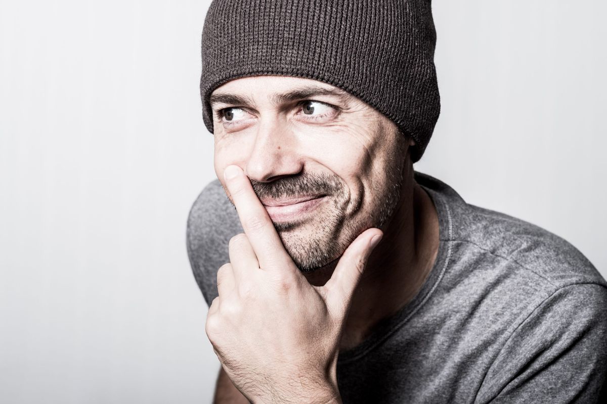 A man in a beanie glancing sideways with a wry, self-aware smile, hand to his chin.