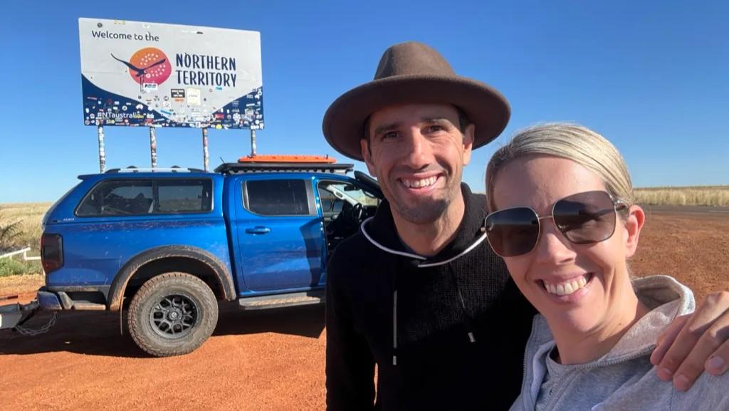 Steph and Matt at the Northern Territory border sign, with their ute parked on the red dirt behind them.
