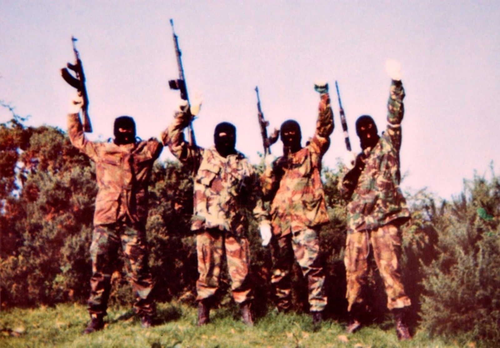 Four masked men in camouflage clothing stand outdoors holding rifles aloft, posing together in a show of armed militancy.