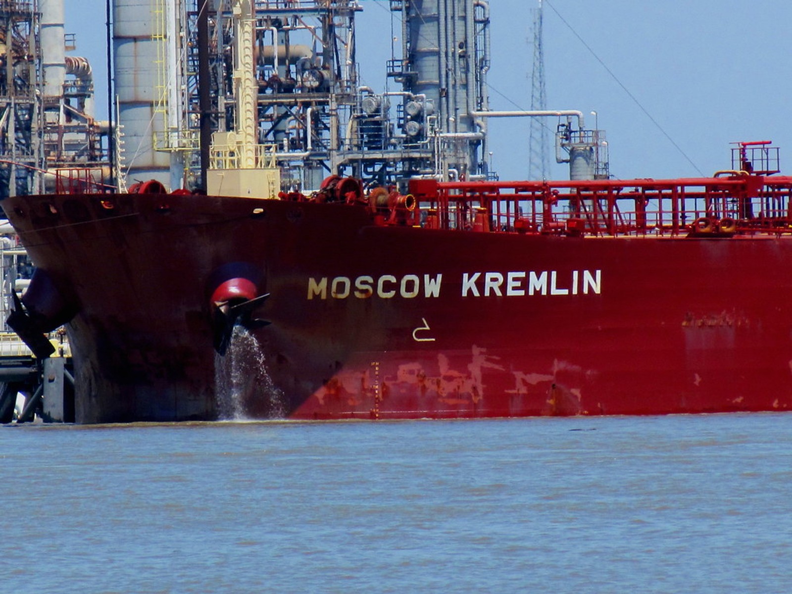 Oil tanker named “Moscow Kremlin” docked at an industrial refinery, with pipes and processing towers visible in the background.