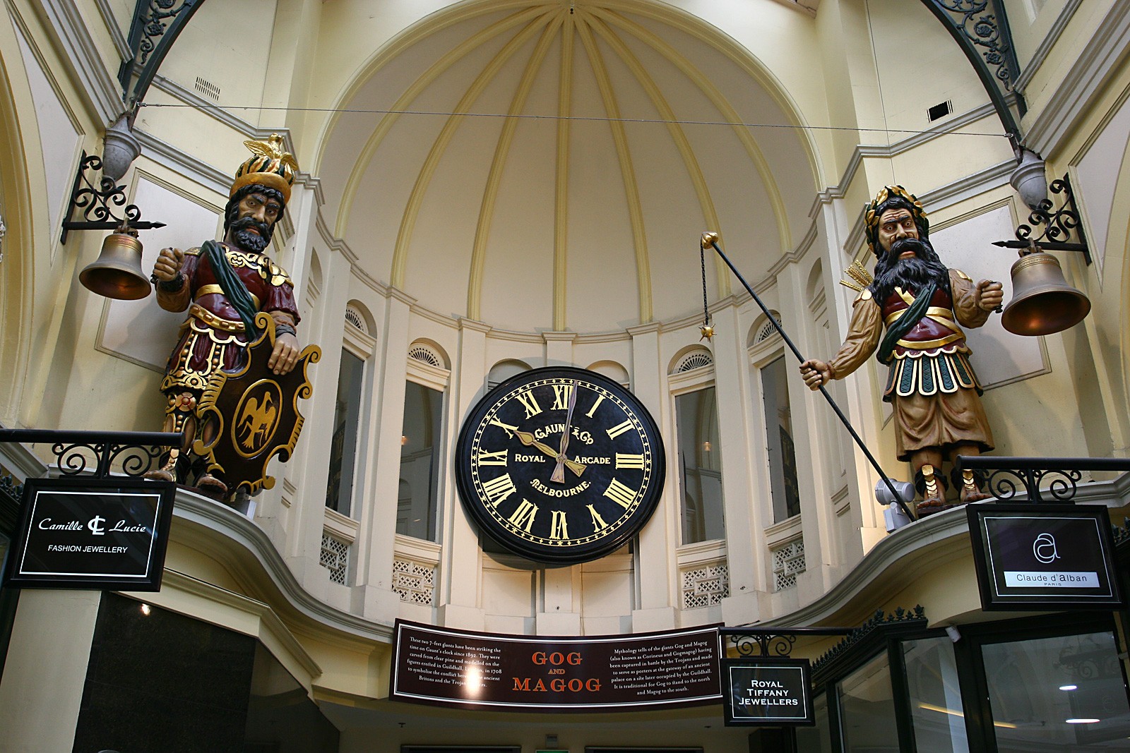 Two large carved statues of Gog and Magog stand on opposite balconies inside Melbourne’s Royal Arcade, flanking a large central clock beneath a high domed ceiling.