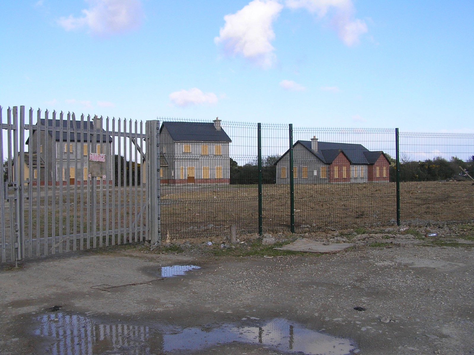 Fenced-off unfinished housing estate with boarded-up homes and empty lots in rural Ireland.
