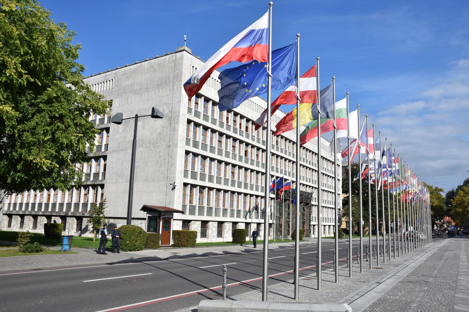 Rows of national flags, including the EU flag, line the street outside the National Assembly building in Ljubljana, with the modernist parliament building visible in the background.