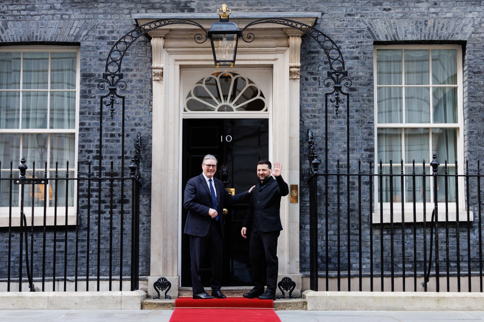 Two political leaders stand outside 10 Downing Street in London, shaking hands on the doorstep, with the iconic black door marked “10” behind them.