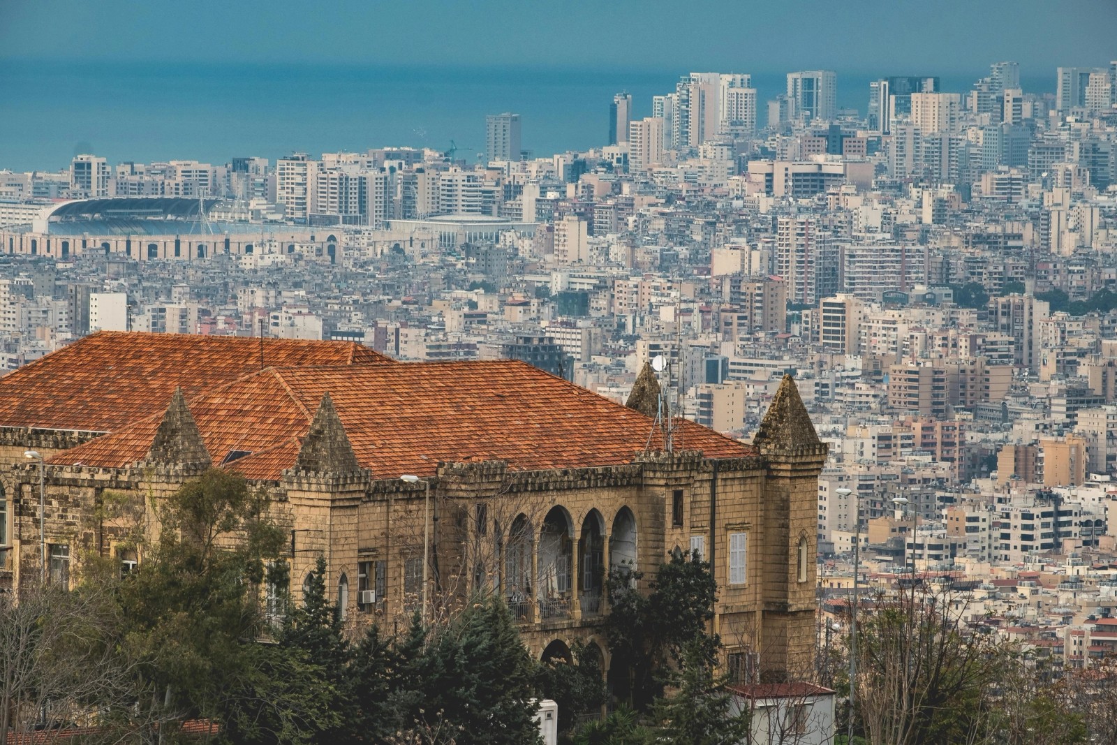 View of Beirut with a historic stone building with a red-tiled roof in the foreground and the dense modern city stretching to the Mediterranean Sea in the background.