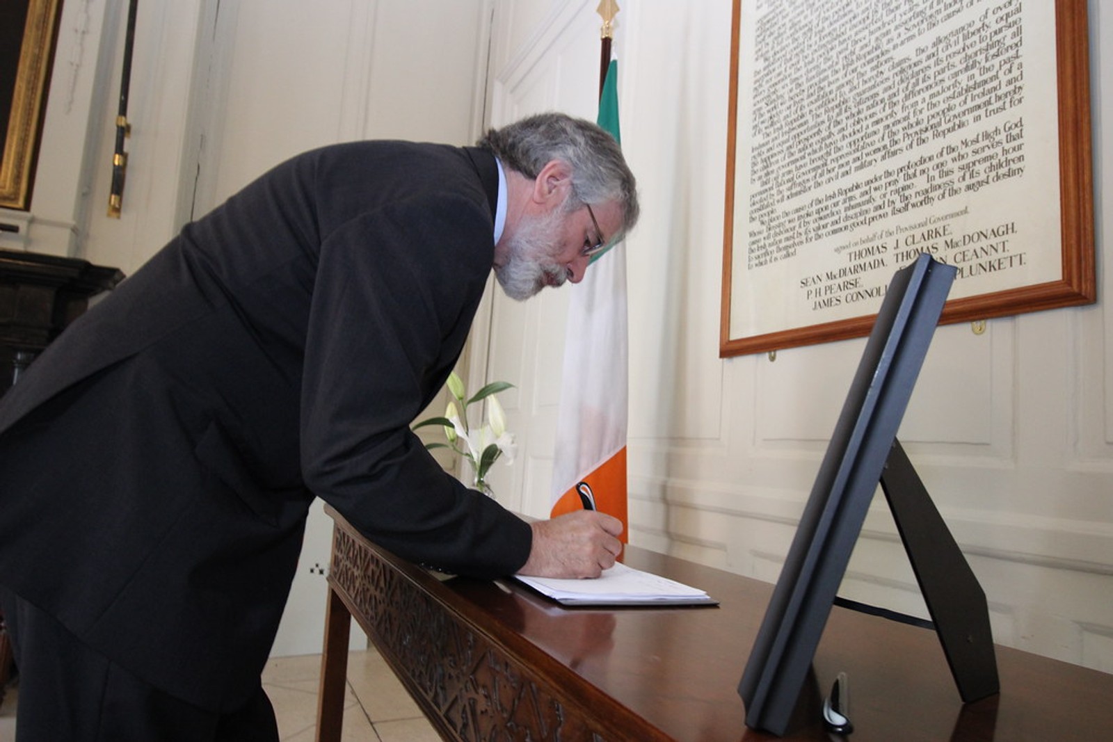 Gerry Adams signing a book of condolences beside an Irish flag and a framed proclamation in a formal room.