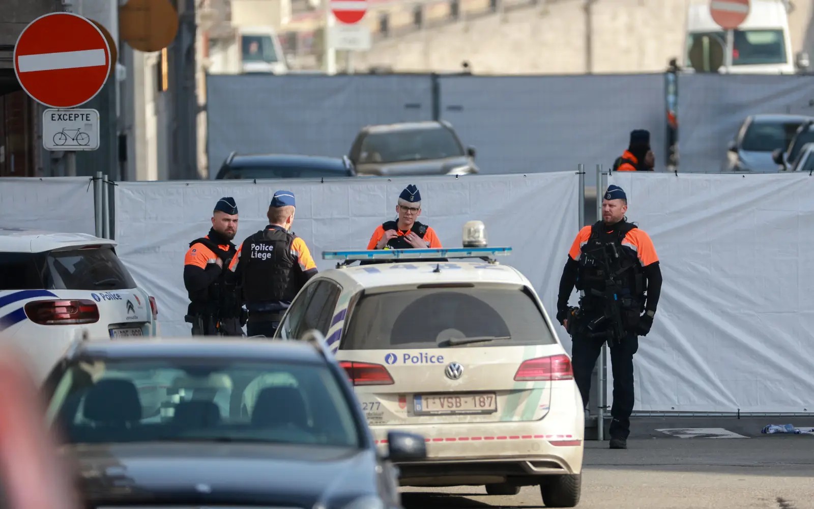 This image shows police officers in Liège, Belgium, standing near police cars on a city street. They are wearing uniforms with orange vests and are behind a white barrier. The scene looks orderly.