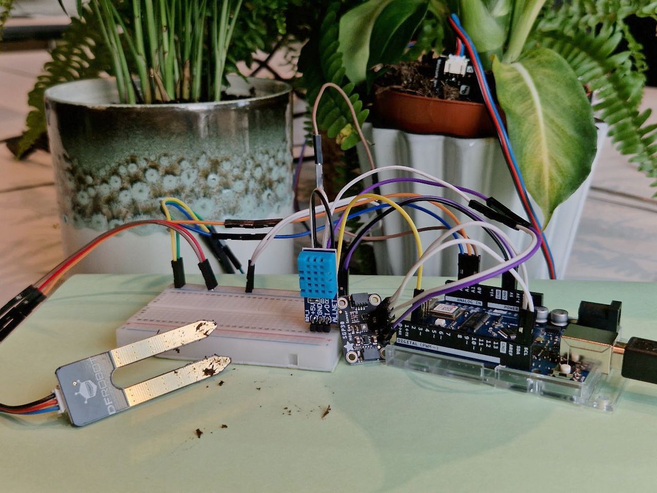 Arduino Komponenten mit verbundenen Breadboard, Bodenfeuchtigkeits-, Luftfeuchtigkeits- und Temperatursensor mit Topfplfanzen im Hintergrund