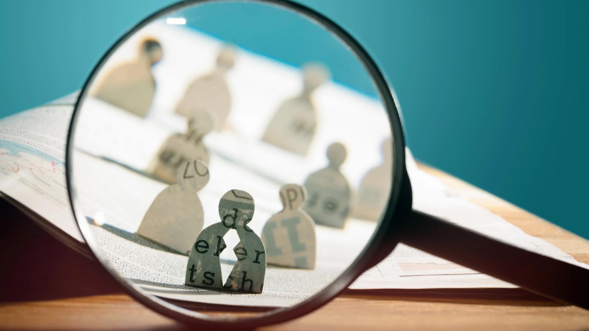 Magnifying glass focusing on paper cut-out figures, symbolizing research and analysis in AI developer recruitment at https://codeautomation.ai/