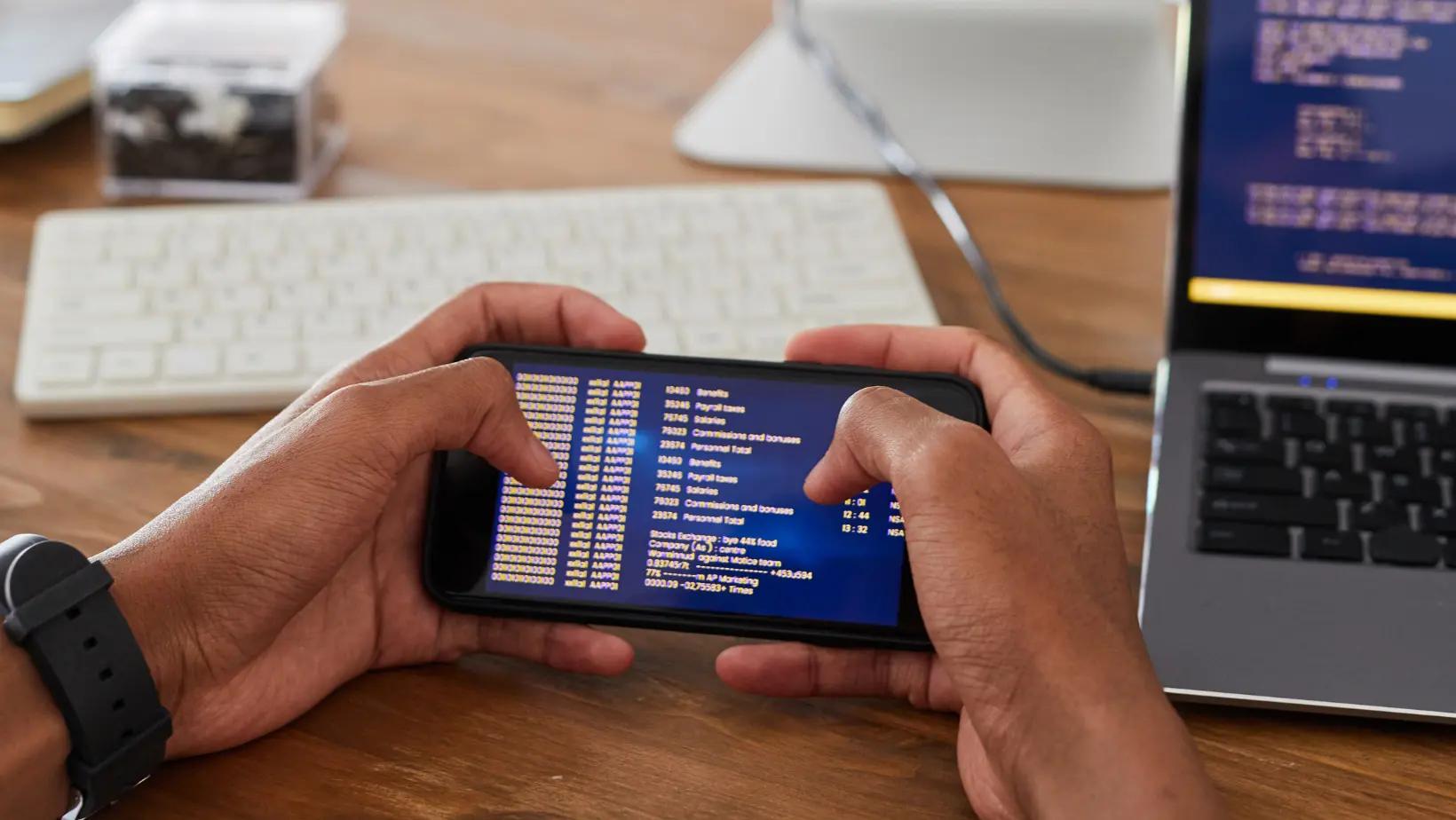 Developer debugging an Android app on a smartphone, with coding scripts visible on a laptop screen