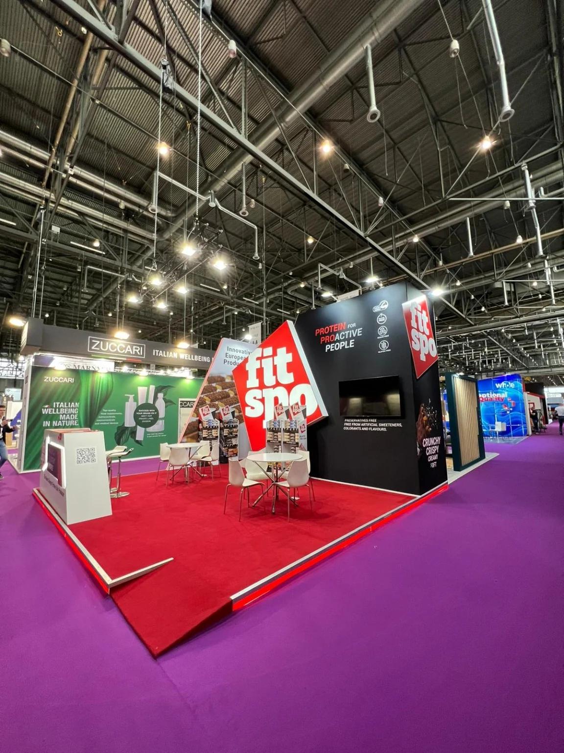 Side view of the FitSpo stand with open access, meeting tables and a screen integrated into the black wall.
