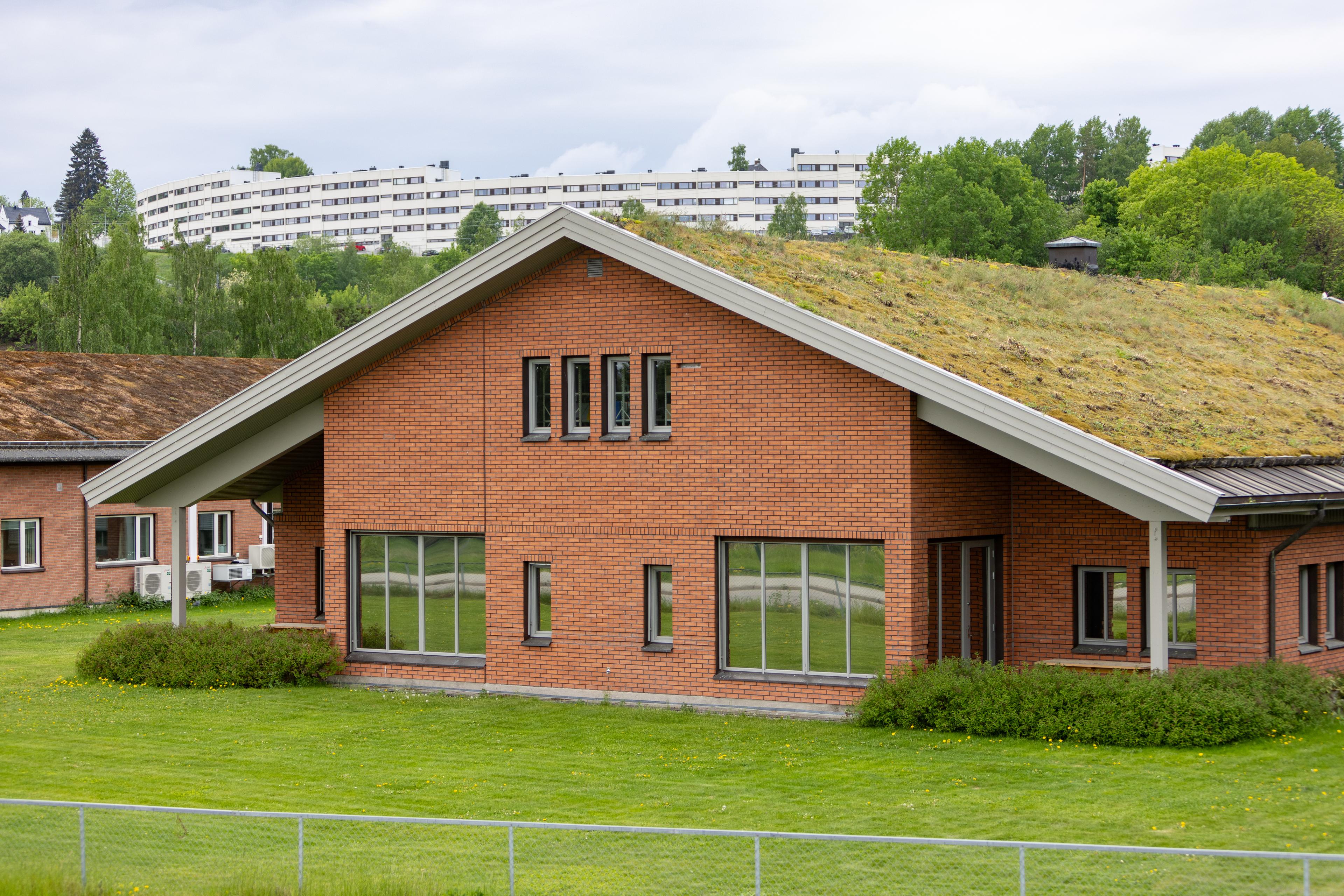 Teglsteinsbygning med grønt tak, omgitt av plen, med boligblokker i bakgrunnen